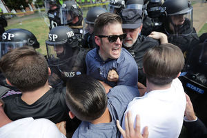 White man in blue shirt surrounded by White men in multicolored clothes and White police officers in black riot gear in front of green grass