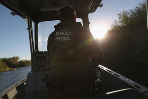 Border Patrol stands and rides small boat near U.S. Mexico border> His back is to the camera and the back of his jacket reads, Border Patrol. 